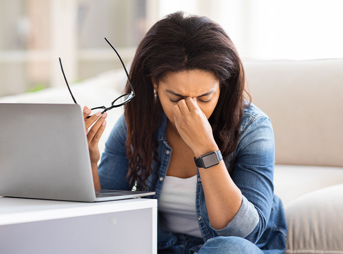 Frustrated woman sitting by laptop, holding glasses and rubbing her eyes, reflecting family drama over grandson’s ashes.