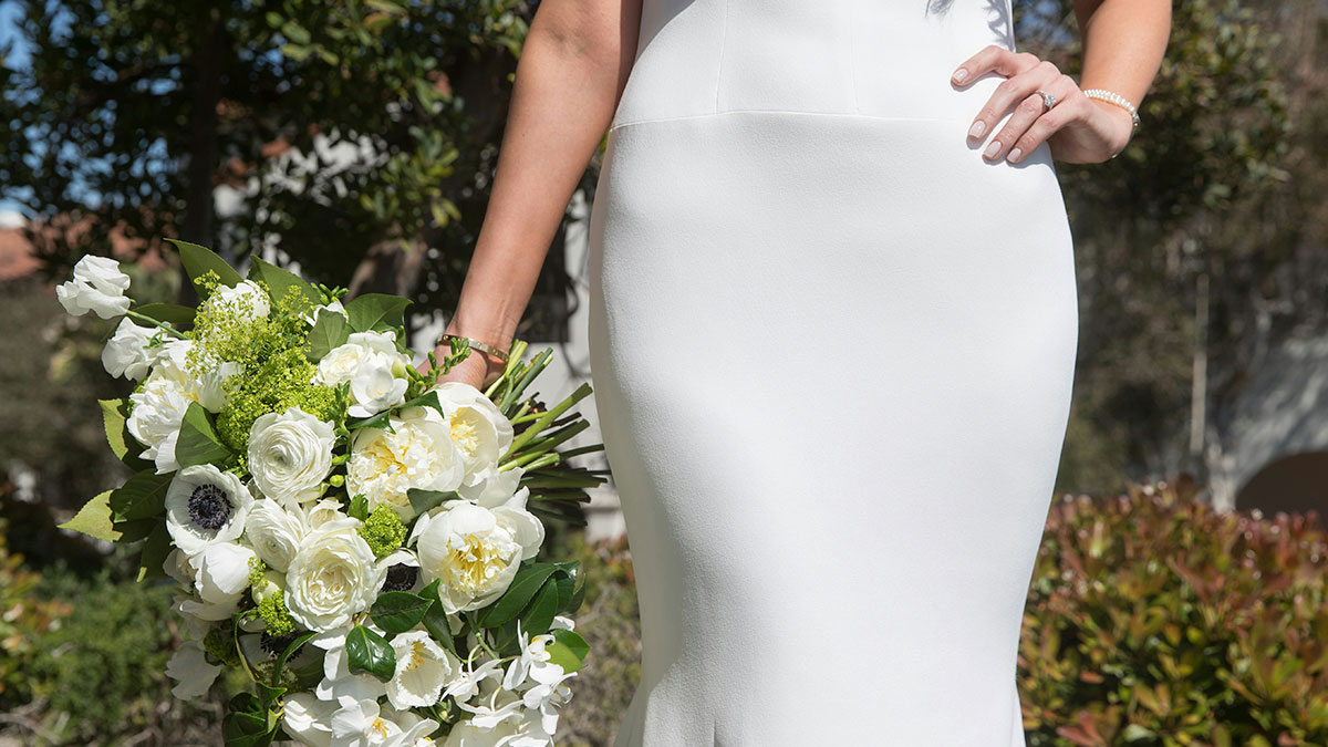 Bride in a fitted white wedding dress holding a bouquet of white flowers in an outdoor garden setting