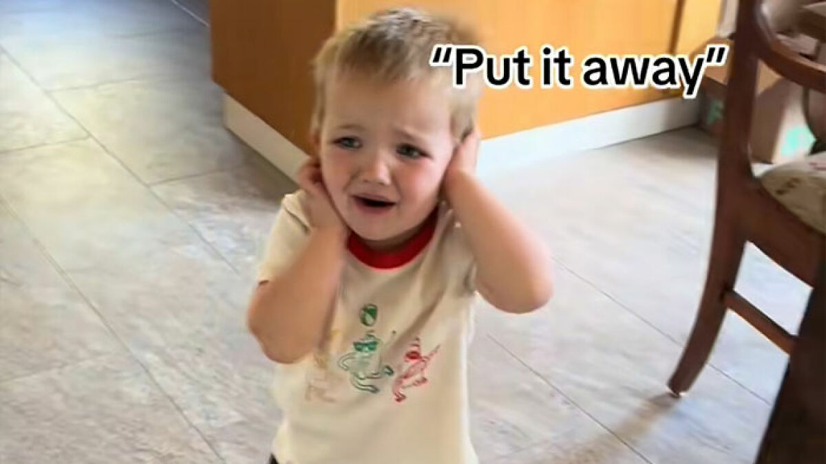 Toddler covering ears and complaining about ultrasound photo noise while standing inside a kitchen area.