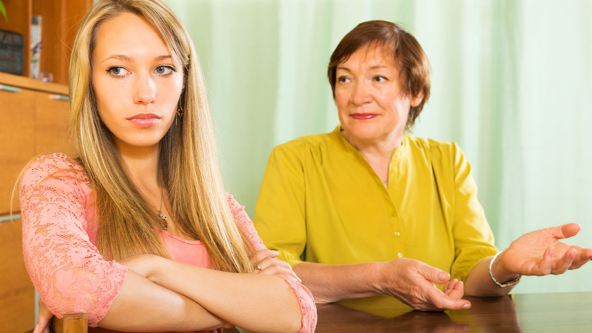 Young woman looking uncomfortable while older woman in yellow discusses baby name and Christian beliefs.