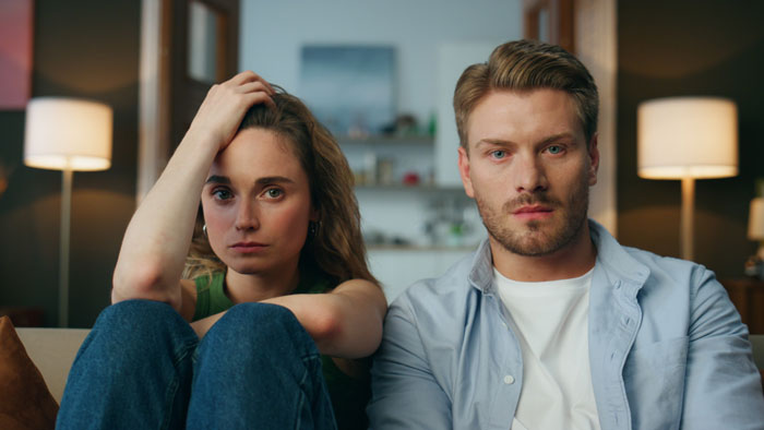 Couple planning a last stress-free trip before wedding, sitting concerned in living room with warm lighting background. Couple planning a last stress-free trip before wedding, sitting concerned in living room with warm lighting background.