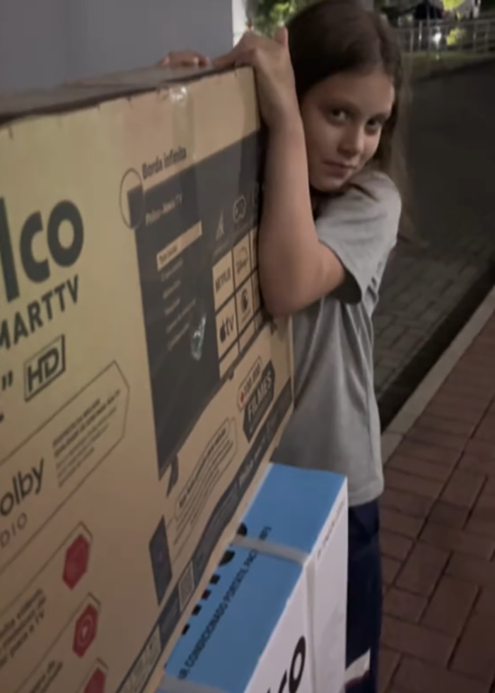 Young girl carries large TV box outdoors, illustrating momfluencer controversy involving autistic daughter and balcony sleeping arrangement. Young girl carries large TV box outdoors, illustrating momfluencer controversy involving autistic daughter and balcony sleeping arrangement.