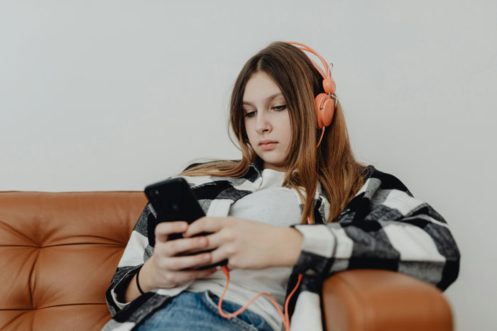 Teen girl with orange headphones sitting on a couch, focused on her smartphone, representing wealthy grandpa gift conflict. Teen girl with orange headphones sitting on a couch, focused on her smartphone, representing wealthy grandpa gift conflict.