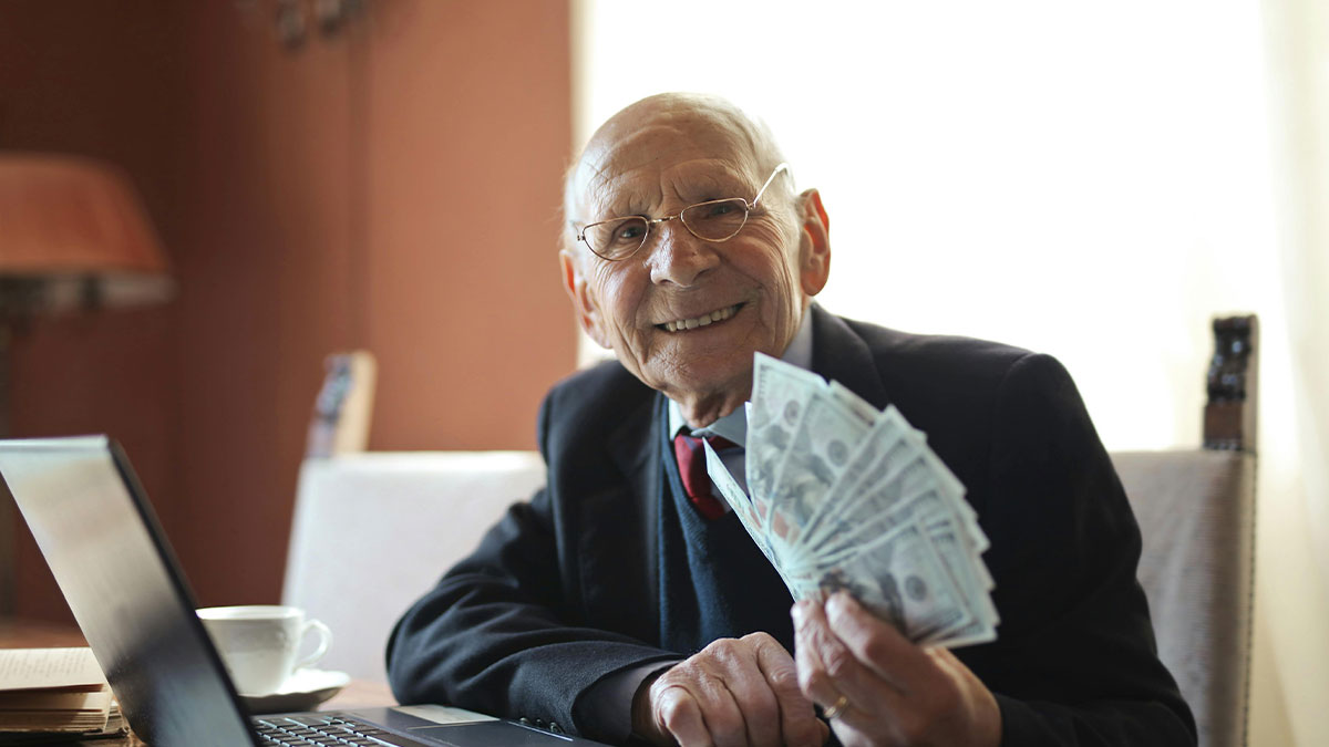 Elderly man smiling while holding cash, sitting at table with laptop, symbolizing wealthy grandpa gifting grandson money.