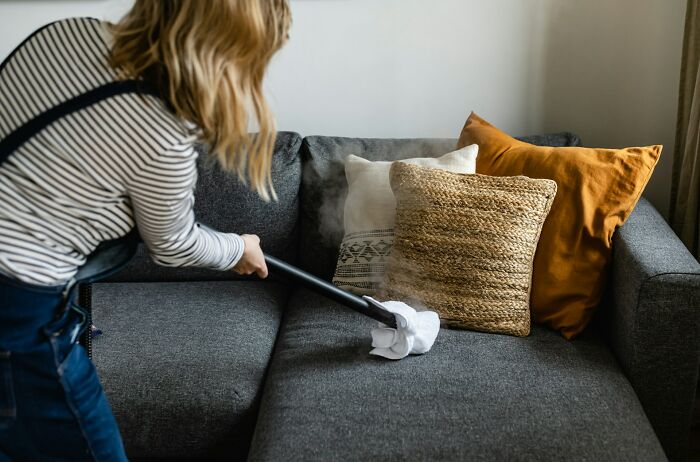 Woman using a steam cleaner on a gray couch with decorative pillows, demonstrating calm and focused communication skills.