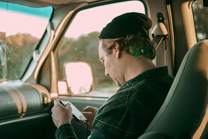 Man wearing a beanie and plaid shirt sitting in a car, looking down at his phone, reflecting on generational trauma discussion.