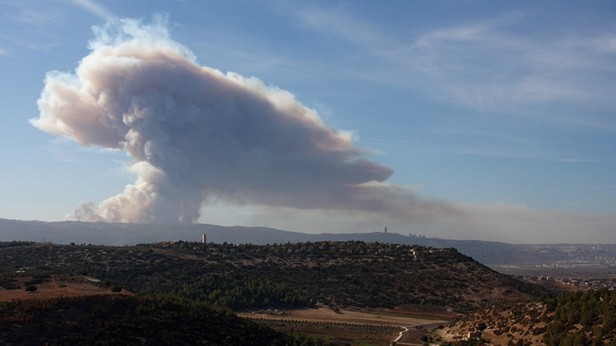 Thick smoke plume rising from a large catastrophic wildfire over a hilly forested landscape under clear sky.