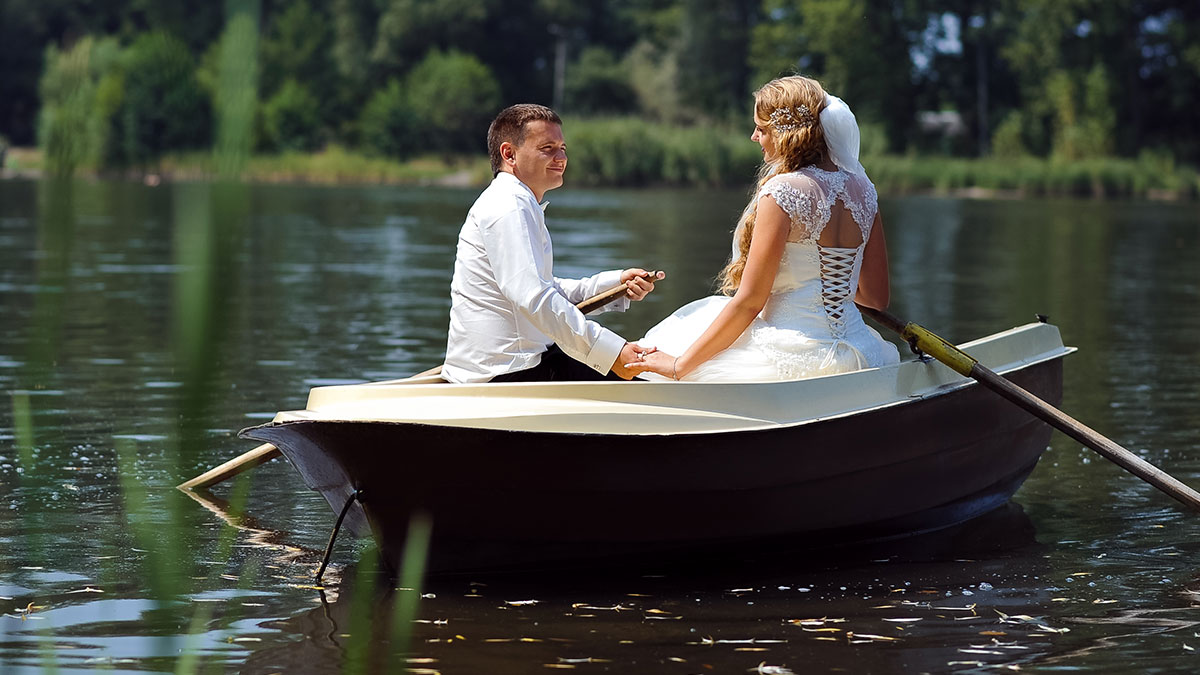 Bride and groom in wedding attire holding hands while sitting in a boat on a calm lake during their wedding day.