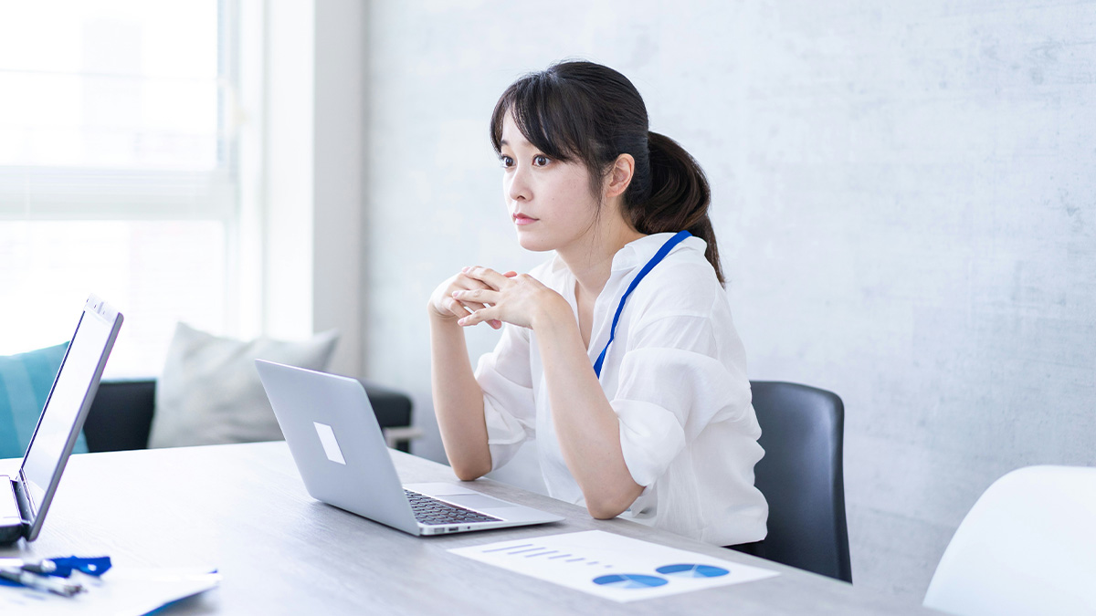 Korean woman in a white shirt sitting at a desk with a laptop, appearing thoughtful and concerned in an office setting