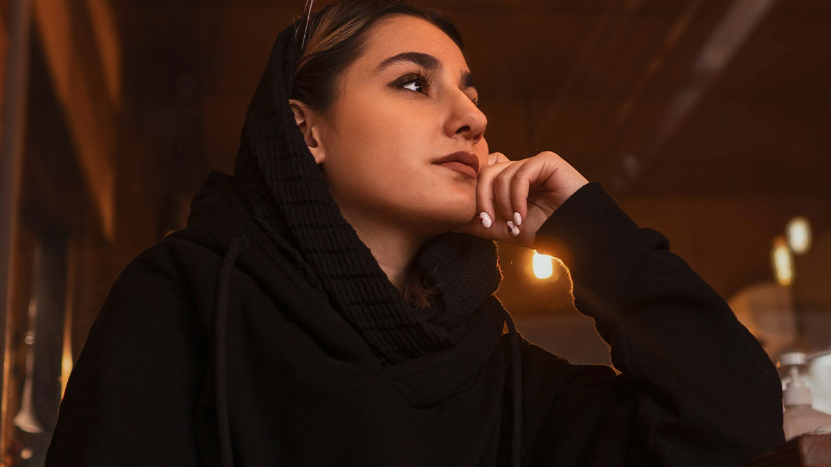 Young Muslim woman wearing black hijab, sitting thoughtfully indoors with warm lighting in the background