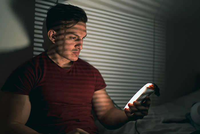 Man looking at phone with shadows of blinds on his face, representing man publicly demoted to friend by GF. Man looking at phone with shadows of blinds on his face, representing man publicly demoted to friend by GF.