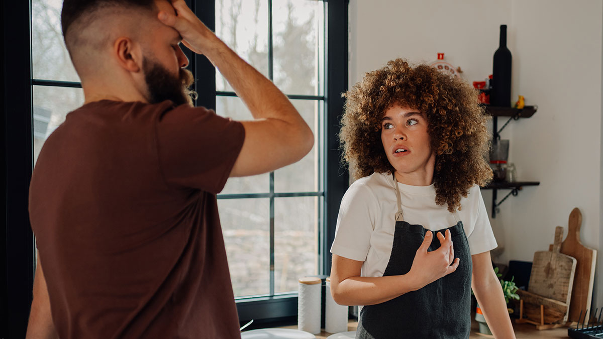 Young woman in apron looks surprised talking to a man in brown shirt about not telling roommate bought house until closed.