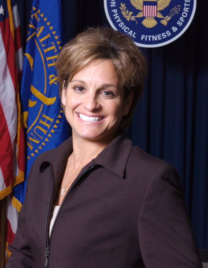 Olympic gold medalist Mary Lou Retton smiling in a professional setting with US flags and an official seal behind her Olympic gold medalist Mary Lou Retton smiling in a professional setting with US flags and an official seal behind her