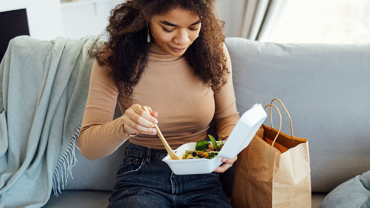 Young woman sitting on a couch eating food from a takeout container, using chopsticks, paper bag beside her, order DoorDash while babysitting.