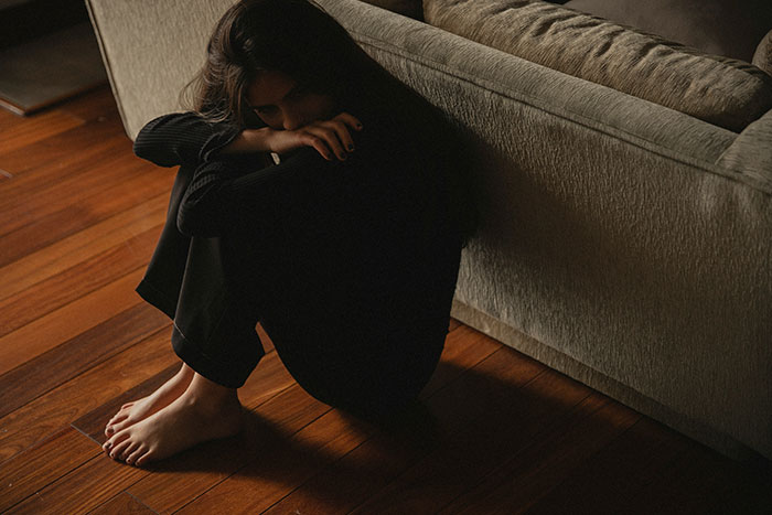 Woman sitting on wooden floor in dark clothing looking down, conveying emotion after man publicly demoted to friend by girlfriend Woman sitting on wooden floor in dark clothing looking down, conveying emotion after man publicly demoted to friend by girlfriend