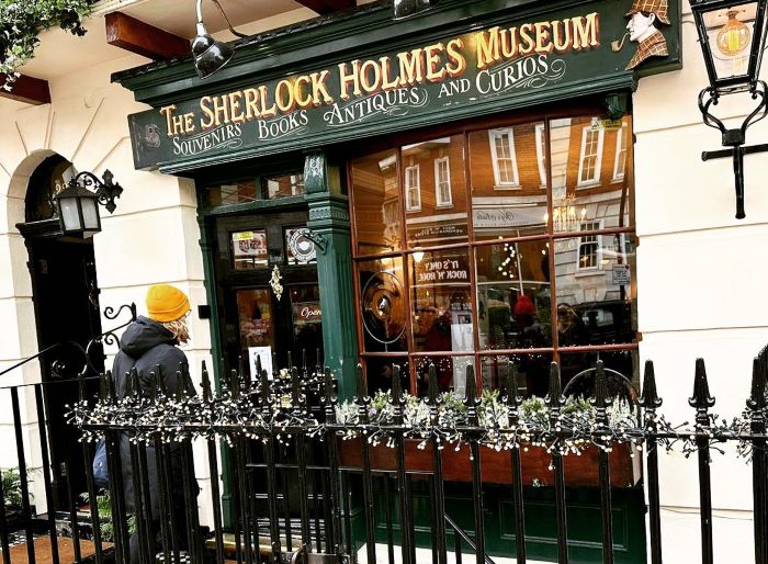 Person in a yellow hat standing outside the Sherlock Holmes Museum amid holiday decorations and reflections in the window. Person in a yellow hat standing outside the Sherlock Holmes Museum amid holiday decorations and reflections in the window.