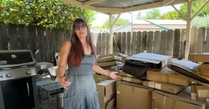 Woman in a sleeveless dress standing by hundreds of mistakenly delivered Amazon packages stacked under a covered patio area. Woman in a sleeveless dress standing by hundreds of mistakenly delivered Amazon packages stacked under a covered patio area.