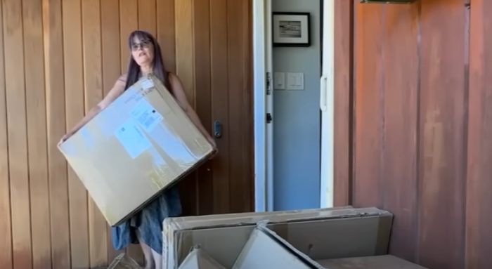 Woman holding large Amazon package at home with multiple mistakenly delivered Amazon packages stacked nearby.