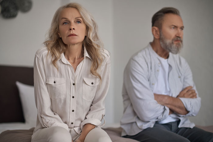Middle-aged parents sitting apart on a bed, looking upset and distant, highlighting challenges in kid care and parenting. Middle-aged parents sitting apart on a bed, looking upset and distant, highlighting challenges in kid care and parenting.
