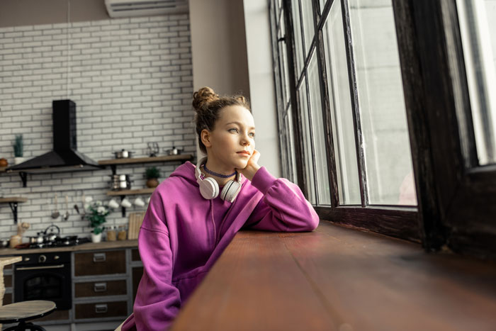 Young woman in a purple hoodie with headphones, sitting by a window, reflecting on leaving toxic parents and shared roof struggles.