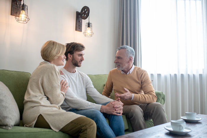 Young son sitting with in-laws on a couch discussing helping with baby while mother appears overprotective nearby. Young son sitting with in-laws on a couch discussing helping with baby while mother appears overprotective nearby.