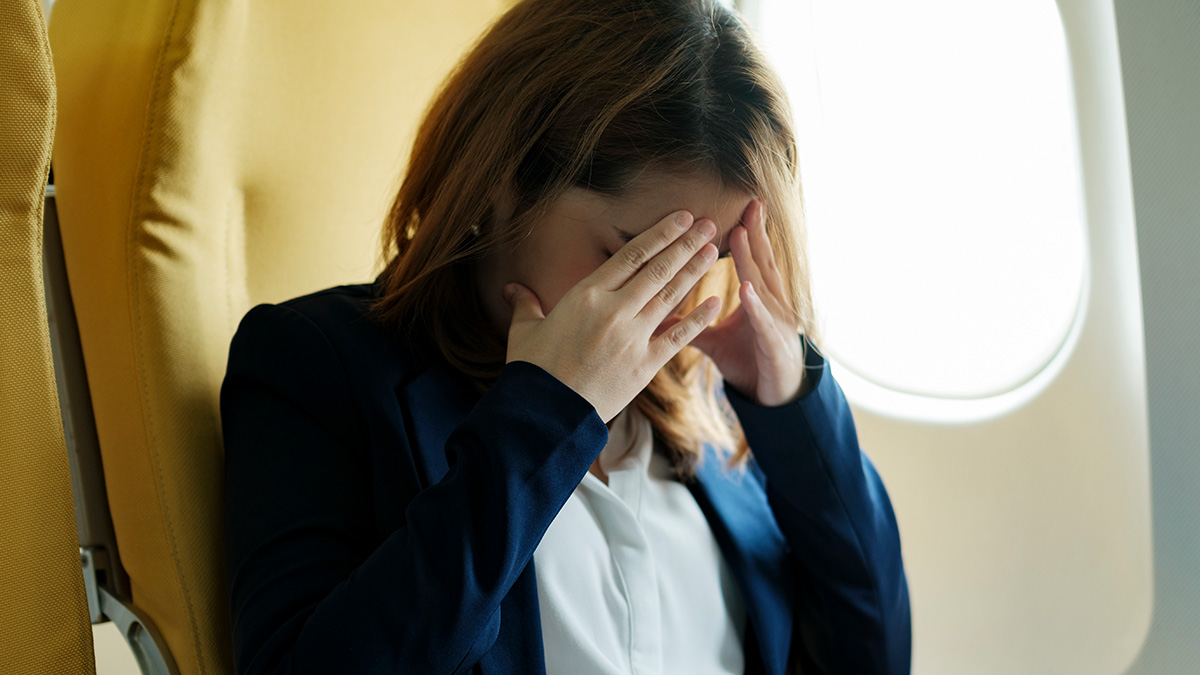 Woman blamed for rowdy kids on flight, sitting stressed in airplane seat by window with hands on her face.