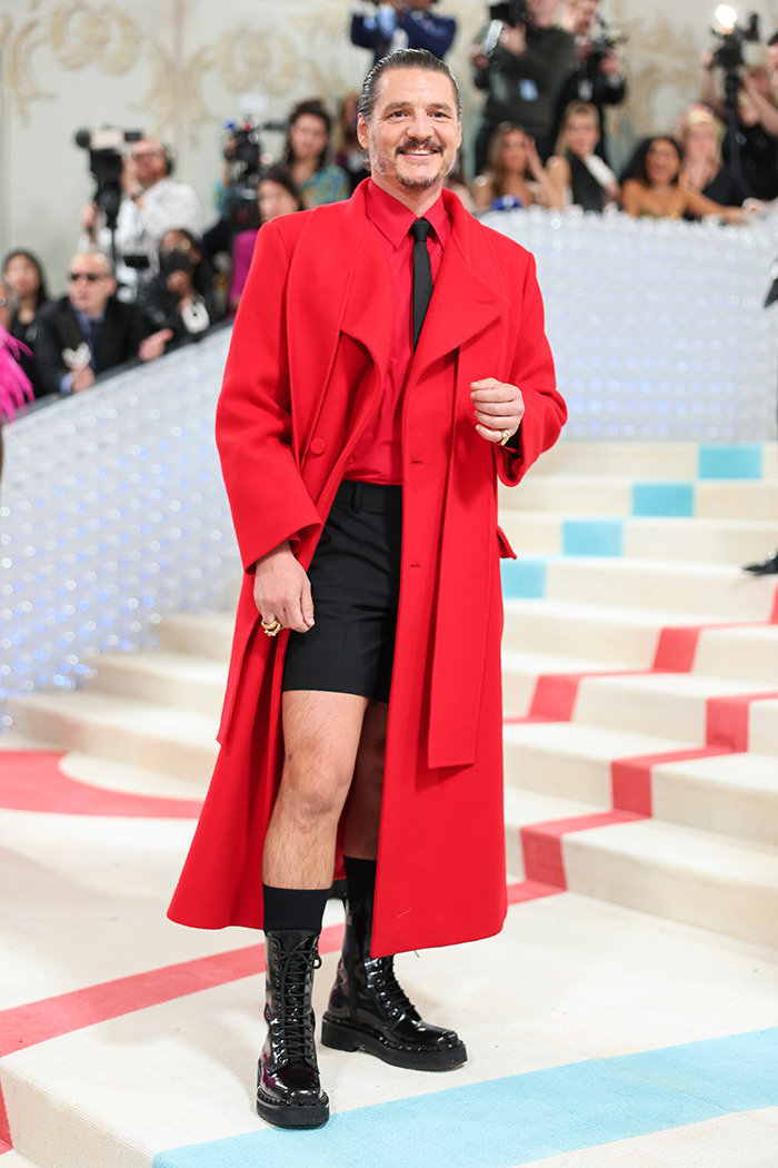 Pedro Pascal wearing a bold red coat and black boots at a high-profile fashion event with photographers in the background Pedro Pascal wearing a bold red coat and black boots at a high-profile fashion event with photographers in the background