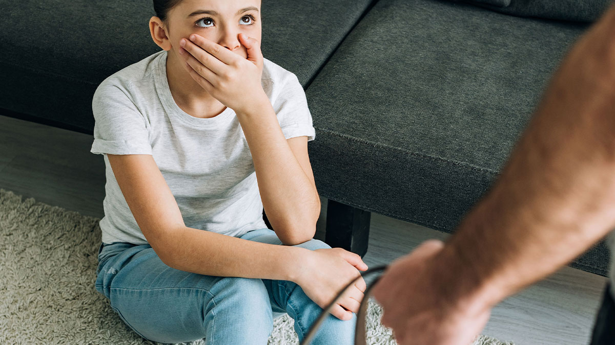 Young person sitting on the floor looking scared while an adult holds a belt nearby in a tense home environment.