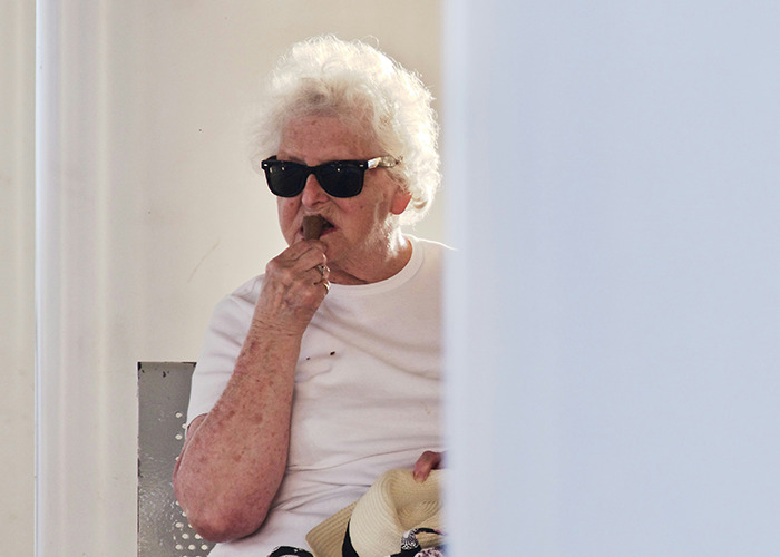 Elderly woman wearing sunglasses and a white shirt, holding a hat and biting her fingernail in a candid moment.