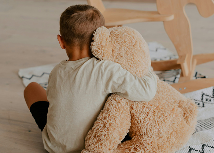 Young boy hugging a large teddy bear on the floor, capturing a moment of comfort amid savage insults shared online.