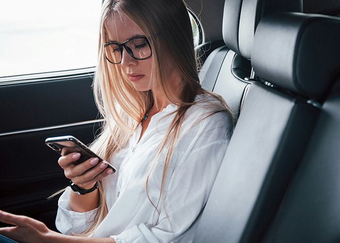 Young woman with glasses sitting in car, focused on her phone, illustrating reactions to savage insults shared online.
