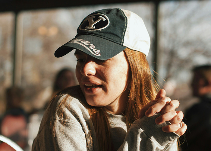 Young woman wearing a cap and sweatshirt, engaging in conversation with a thoughtful expression and folded hands.