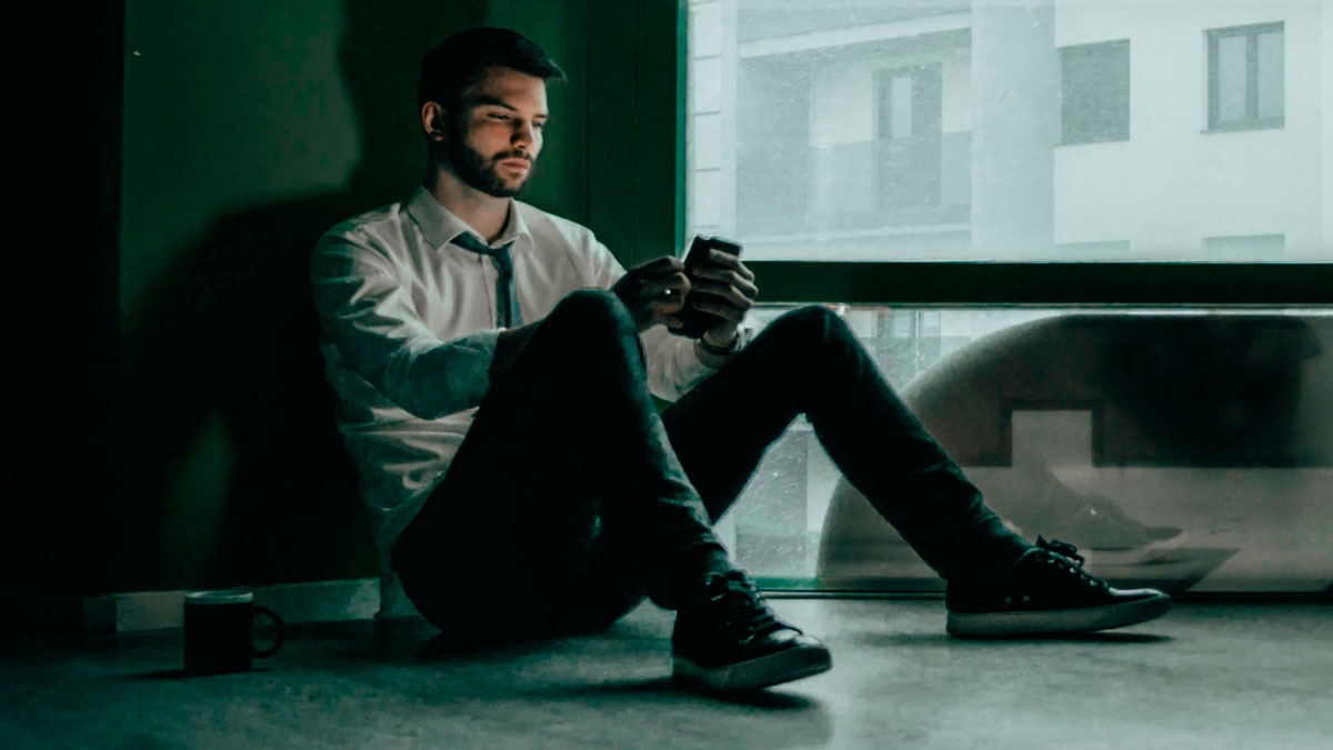 Man in a white shirt sitting on the floor by a window, looking sad and reflecting on brutal truths about life.