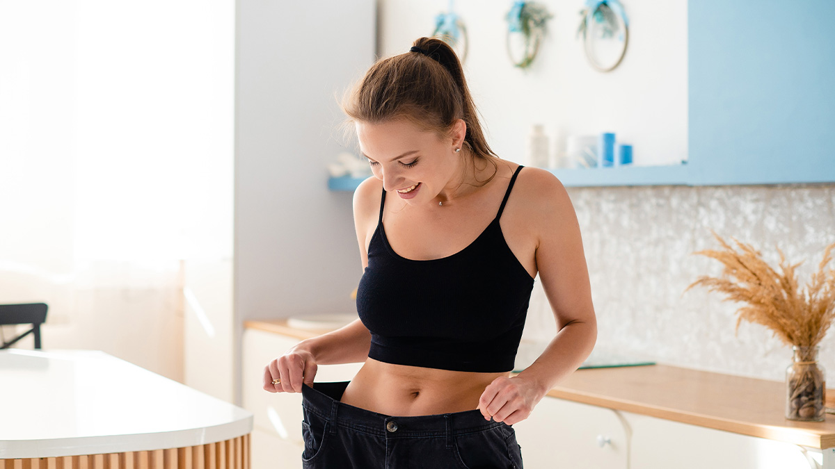 Woman in a black top smiling while holding loose pants, illustrating impact of loved ones taking Ozempic on weight loss.