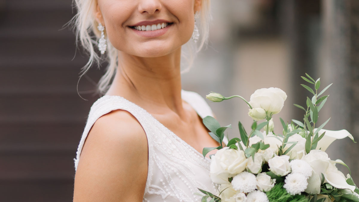 Smiling woman in a white dress holding a bouquet of white flowers, representing family and personal stories.