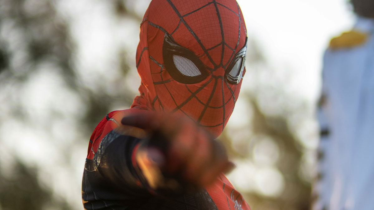 Person dressed in a Spider-Man costume pointing, with blurred trees and a person in white uniform in the background.