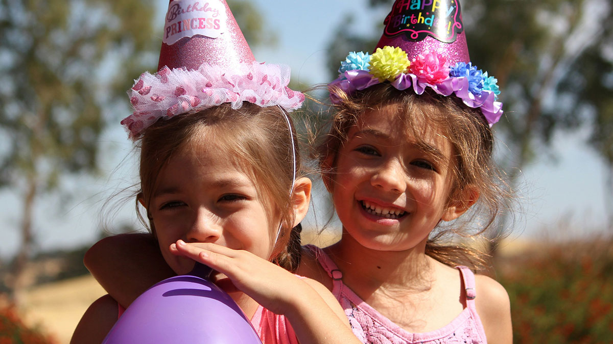 Two young girls wearing birthday hats smiling outdoors holding a purple balloon on a sunny day.