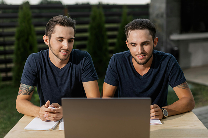 Twin gay men sitting outdoors at a table with a laptop, wearing navy shirts, discussing plans with partner. Twin gay men sitting outdoors at a table with a laptop, wearing navy shirts, discussing plans with partner.