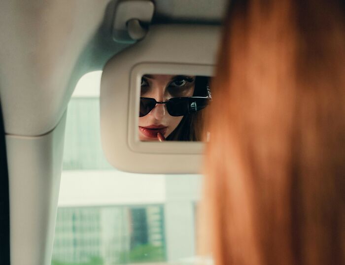 Woman with sunglasses applying lipstick in a truck mirror, capturing truckers share craziest things seen moment.