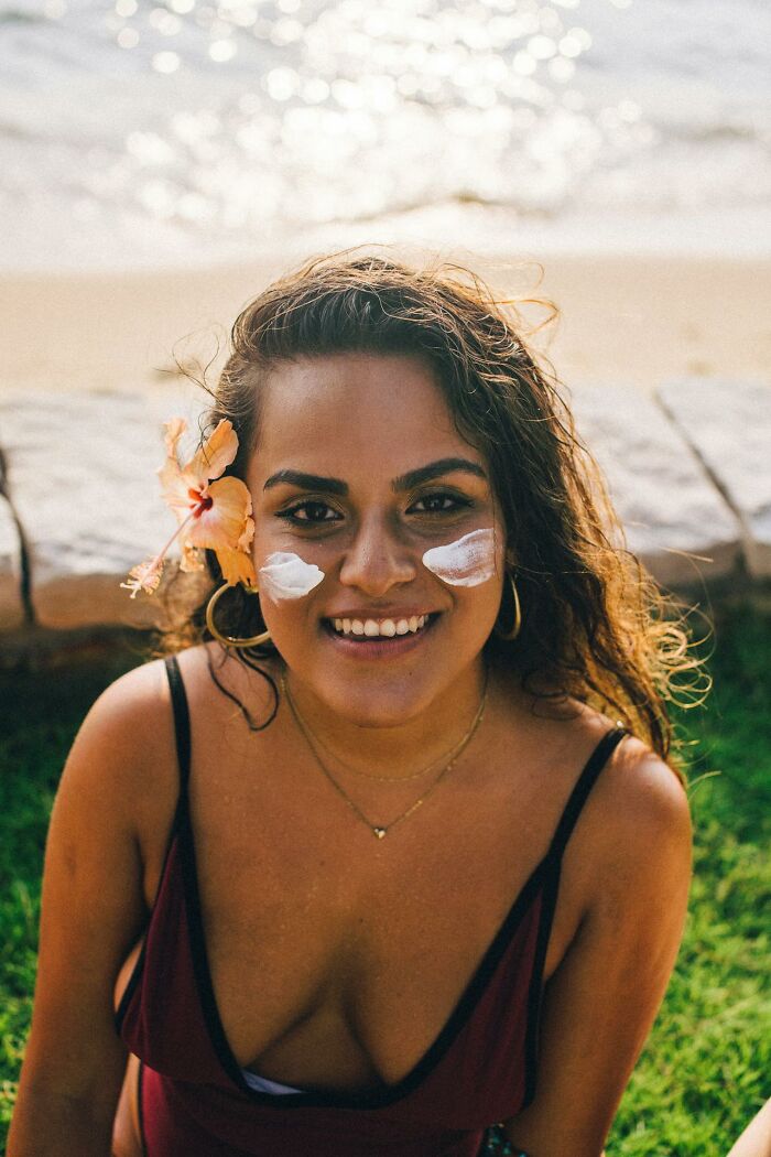 Young woman at the beach with sunscreen on cheeks, wearing flower in hair, smiling and promoting stay hydrated lifestyle.