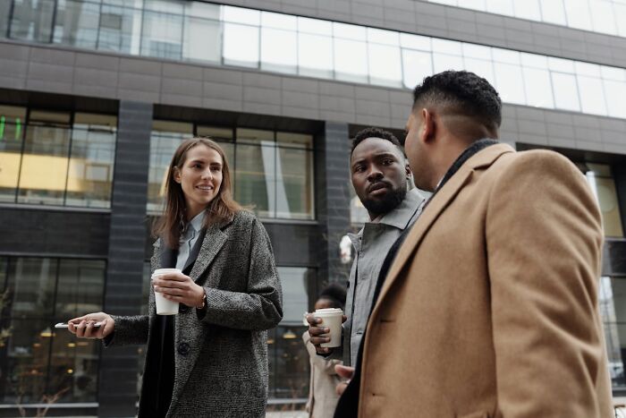Three professionals having coffee outside a modern office building, discussing ways to take revenge on their bosses.