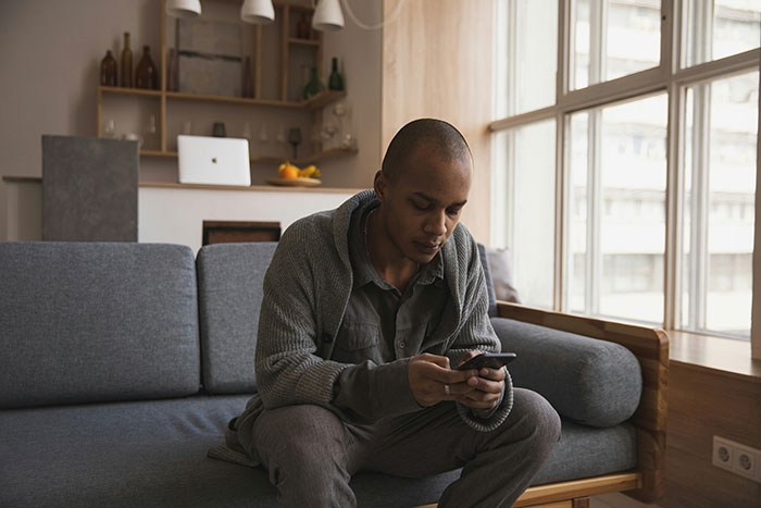 Man sitting on gray couch using phone indoors, depicting tension related to fiancé’s female friend demanding speech and best man role. Man sitting on gray couch using phone indoors, depicting tension related to fiancé’s female friend demanding speech and best man role.