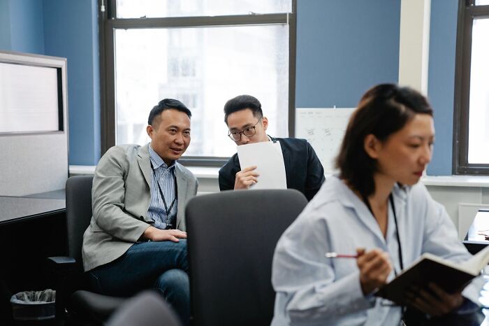 Two office workers whispering and sharing papers near coworkers in a modern workspace, hinting at revenge on bosses.