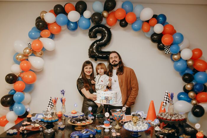 Family celebrating child's second birthday with colorful balloons and toy cars, reflecting trends leading to overconsumption.