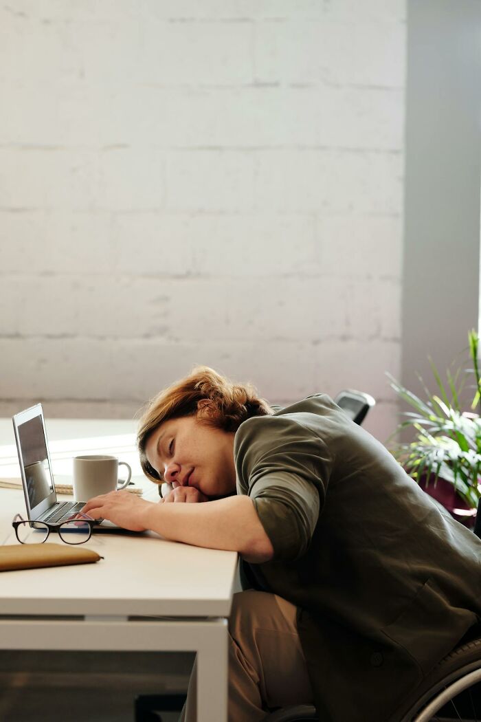 Person exhausted at desk leaning on laptop with coffee cup nearby, symbolizing revenge on bosses through workplace frustration.