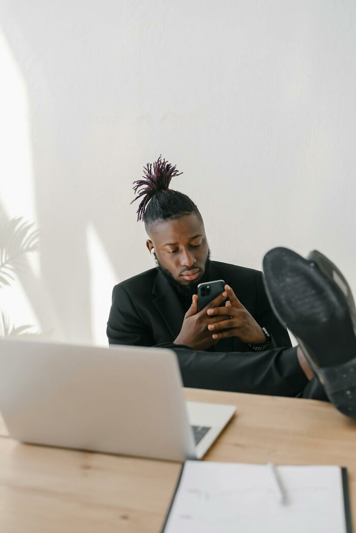 Young man in a black suit sitting at desk with laptop, using phone, illustrating people sharing how they took revenge on bosses.