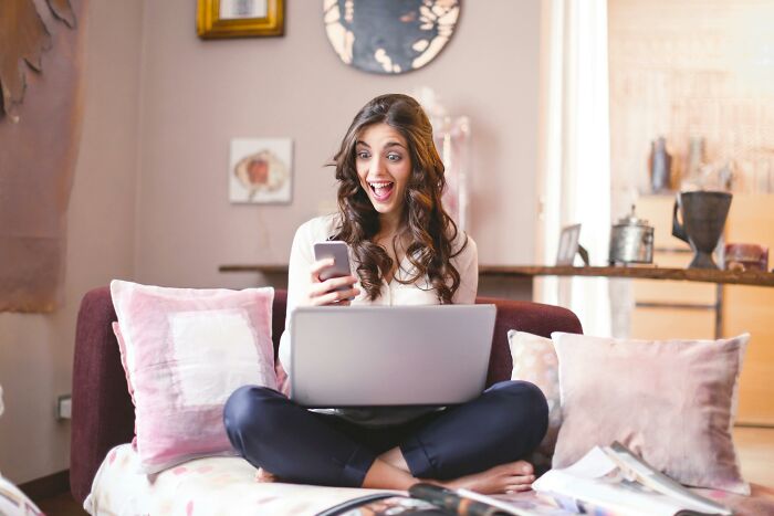 Young woman excitedly taking a Hollywood movie quiz on her phone and laptop while sitting comfortably on a couch. Young woman excitedly taking a Hollywood movie quiz on her phone and laptop while sitting comfortably on a couch.