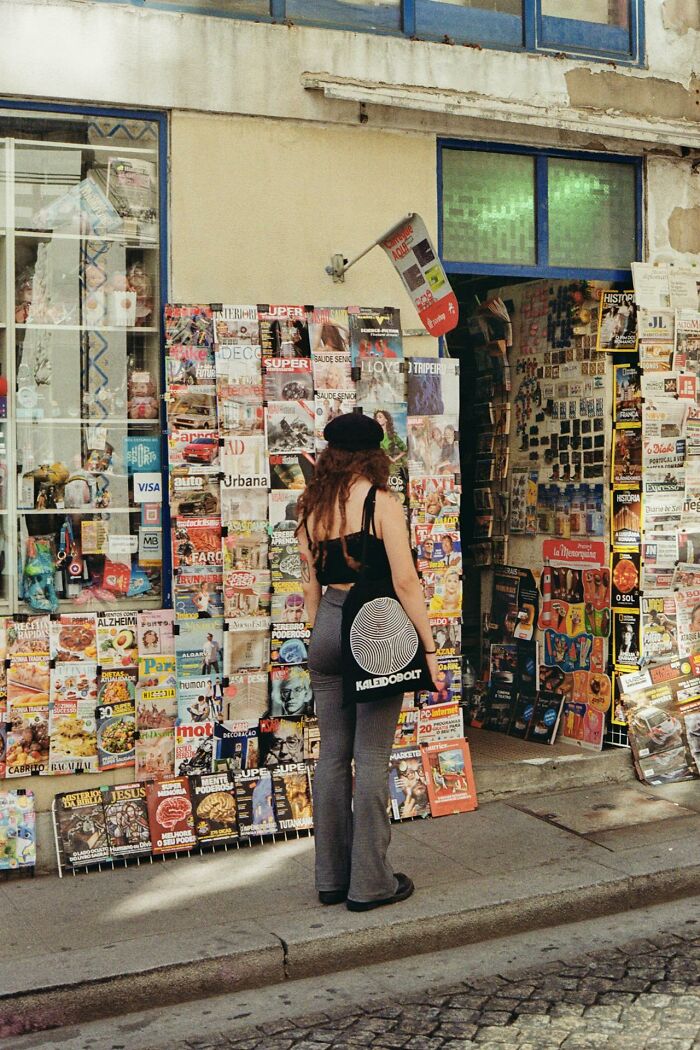 Person with a black beret and tote bag outside a newsstand covered with magazines about revenge on bosses.