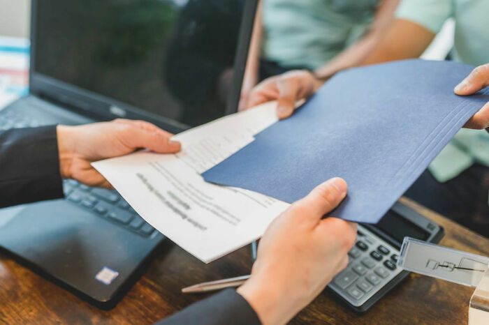 Hands exchanging documents over a desk with a laptop and calculator, illustrating people taking revenge on their bosses.