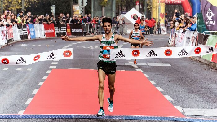 Runner crossing marathon finish line on red carpet with crowd cheering, symbolizing trends leading to overconsumption.